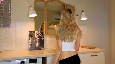 Media: Video of a blonde woman in a white tank top and black shorts, looking at her reflection in a vintage-style mirror with three sections, in a beige-tiled bathroom.