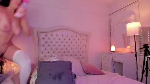 Media: Video of a dimly lit, luxurious bedroom with a tufted headboard, plush pillows, white furniture, and a cozy, glowing lamp.