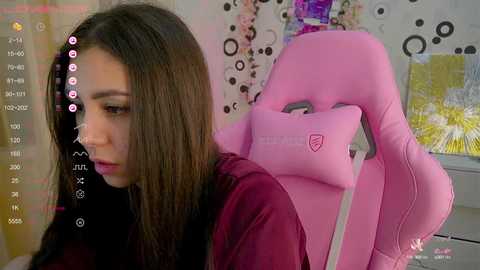 Media: Video of a young woman with long brown hair, wearing a pink gaming chair, in a colorful, cluttered room with a white wall and digital clock displaying 1:23.