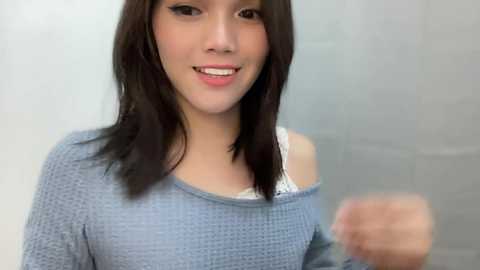 Media: Video of a young Asian woman with straight, shoulder-length dark hair, smiling, wearing a blue off-shoulder knit sweater.