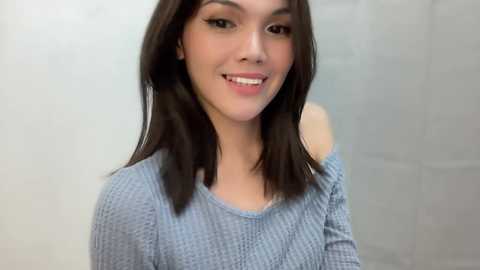 Media: Video of a smiling, light-skinned young woman with straight, shoulder-length dark brown hair, wearing a light blue, off-the-shoulder knitted sweater. Background is a plain, light gray wall.