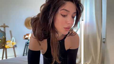 Media: Video of a young woman with long brown hair and fair skin, wearing a black top, leaning forward, indoors with a blurred background.