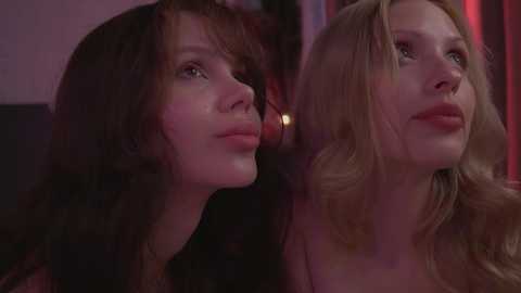 Media: Video of two women with long, wavy hair, one brunette and one blonde, gazing upwards with serene expressions. The background features a dimly lit, red-hued setting.