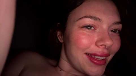 Media: A close-up video of a smiling woman with fair skin, dark hair, and a septum piercing. Her makeup is minimal, highlighting her natural features. The background is dark, focusing attention on her face.