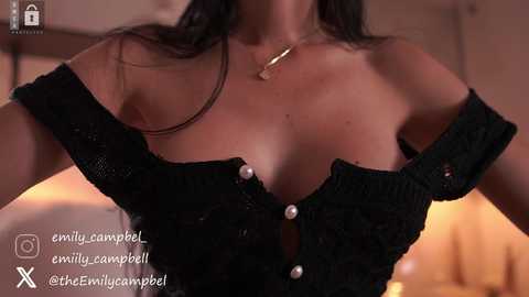 Media: Video of a woman with medium brown skin, wearing a black off-the-shoulder knit top with buttons, and a gold necklace, indoors.