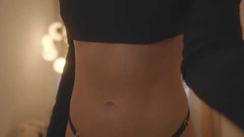 Media: A close-up video of a slender, light-skinned woman's midriff, wearing black lingerie. Her stomach is flat with a small navel. The background is softly lit with a blurred, warm ambiance.