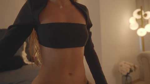 Media: Video of a woman in a black strapless bandeau top, long sleeves, and black pants, indoors with a warm, dimly lit ambiance, featuring a wall-mounted lamp and a bed with a pillow.