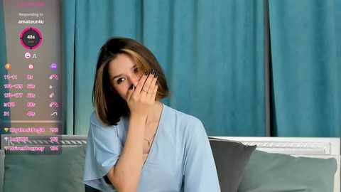 Media: Video of a young woman with shoulder-length brown hair, wearing a light blue hospital gown, covering her mouth with her hand, in a hospital room with teal curtains and medical equipment.