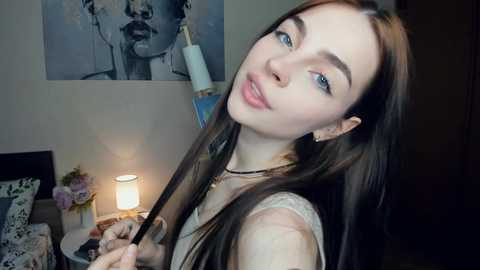 Media: Video of a fair-skinned, long-haired young woman with blue eyes, holding a selfie stick, in a softly lit bedroom with a monochrome art print and a lit candle on a nightstand.