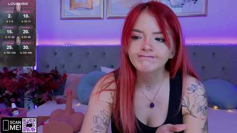 Media: A video of a young woman with vibrant red hair and multiple tattoos, wearing a black tank top, smiling while seated at a table with a wine bottle and glass, in a cozy room with purple lighting.