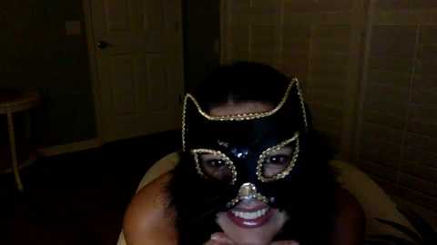 Media: Video of a woman in a black, gold-sequined bat mask, grinning, in a dimly lit room with patterned wallpaper and wooden floor.