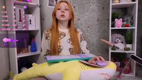 Media: Video of a young woman with long orange hair, wearing a white bear-patterned sweater and yellow leggings, typing on a rainbow keyboard in a room with pink and gray decor.