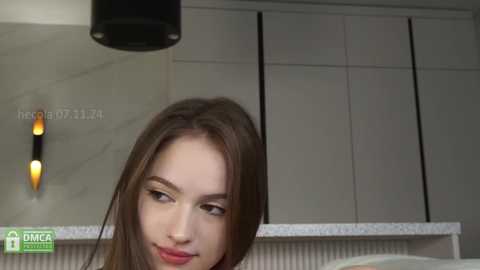 Media: Video of a young Caucasian woman with straight brown hair and fair skin, smiling, in a modern kitchen with white cabinets and a marble backsplash.