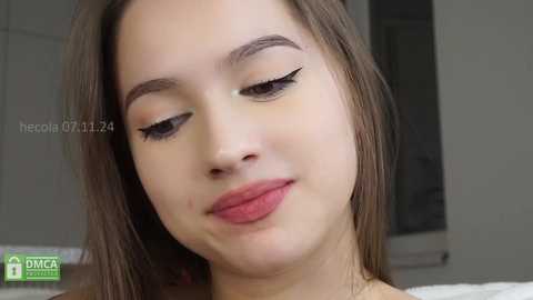 Media: A close-up video of a young woman with fair skin, brown hair, and light makeup, including subtle eyeshadow and winged eyeliner. She has a neutral expression.