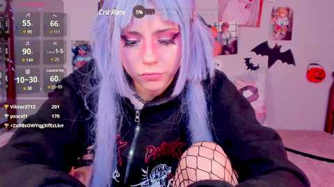 Media: Video of a pale-skinned woman with long, pastel blue hair and black eyeliner, wearing a black jacket with a skull graphic, fishnet stockings, and a distressed expression. The background features a pink room with cartoon posters and a bat decoration.