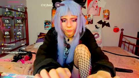 Media: Video of a pale, blue-haired woman with gothic makeup, wearing black clothes and fishnet tights, sitting on a bed in a cluttered room with posters, a gaming controller, and a cat.