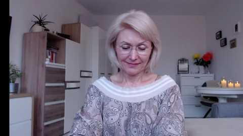 Media: Video of a middle-aged woman with short blonde hair, wearing a patterned, off-the-shoulder top, standing in a modern, light-colored living room with white cabinets, a round table, and candles.