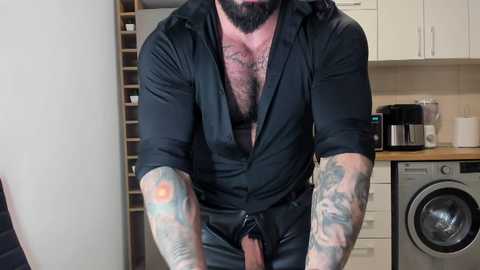 Media: Video of a muscular man with a beard and tattoos, wearing a black unbuttoned shirt, standing in a modern kitchen.