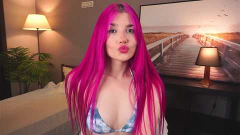 Media: Video of a young woman with long, vibrant pink hair, wearing a tie-dye bikini top, puckering her lips, indoors with a lamp, green plant, and beach-themed artwork in the background.