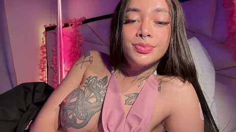 Media: Video of a young, attractive Latina woman with long, straight black hair, wearing a pink halter top, showcasing tattoos on her arms and chest, lying on a bed with pink and white bedding.