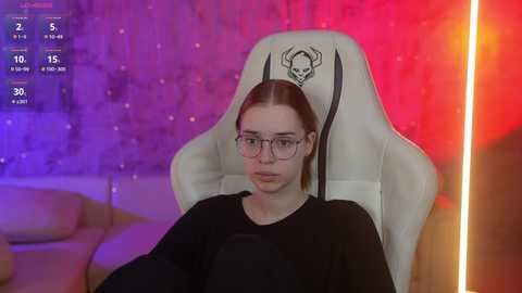 Media: A video of a young woman with glasses, pale skin, and straight brown hair, sitting in a white gaming chair with a skull emblem. The background has a red and purple gradient with a blurred sofa.