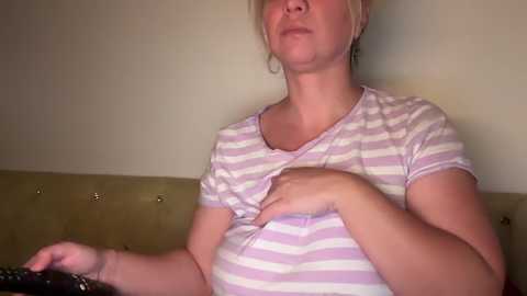 Media: Video of a pregnant woman with fair skin and blonde hair, wearing a light purple and white striped t-shirt, holding a remote control, sitting on a beige couch against a plain beige wall.