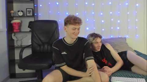 Media: Video of two young, slender, white men with fair skin and red hair, wearing black athletic shorts and shirts, sitting on a bed with blue string lights in the background.