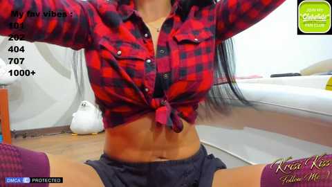 Media: Video of a woman in a red plaid shirt tied at the waist, revealing her toned midriff, wearing purple shorts, indoors. Background includes a white couch and a white statue.