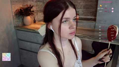 Media: Video of a young, fair-skinned woman with long dark hair, wearing white lace lingerie and white earbuds, holding a red lollipop. She stands in a modern kitchen with a stone countertop and potted plants.