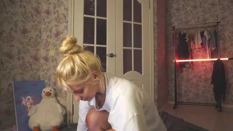 Media: A video of a blonde woman in a white top, kneeling on a dark floor, with a stuffed penguin beside her. The room has floral wallpaper and a wardrobe with clothes hanging inside.