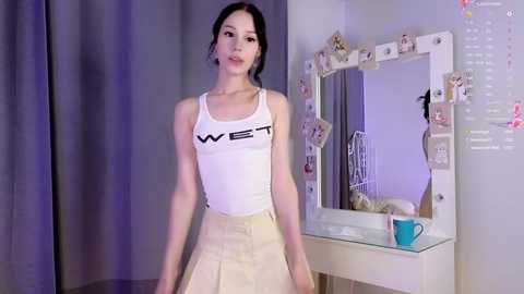 Media: Video of a slender Asian woman with dark hair in a white tank top and beige skirt, standing in a modern bedroom with a mirror and photos on the wall.