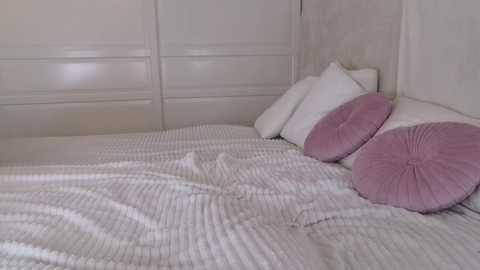 Media: A video of a minimalist bedroom with a white, textured comforter, two white pillows, and a pink, ruffled pillow, all on a white bed against a white wall.