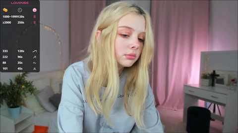 Media: Video of a blonde, fair-skinned woman with light makeup, wearing a light blue sweater, sitting at a desk in a room with pink curtains and a white vanity.