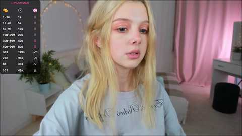 Media: A video of a young, fair-skinned, blonde woman with light pink eyeshadow, wearing a gray sweatshirt, sitting indoors. The room has pink curtains, a white desk, and a potted plant.