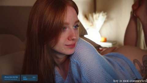 Media: Video of a fair-skinned, red-haired woman in a light blue sweater, lying on a bed in a dimly lit room, with a blurred background featuring a bedside table and lamp.