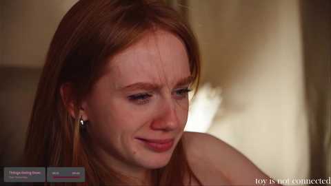 Media: Video of a fair-skinned woman with long red hair, wearing a white top, smiling softly, blurred background.