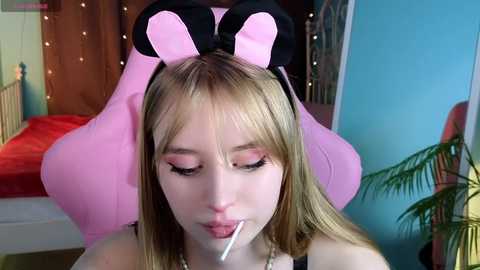 Media: Video of a young, fair-skinned girl with blonde hair wearing a pink Minnie Mouse headband, pink eyeshadow, and sucking on a lollipop. She's indoors with a blue wall, potted plant, and a bed in the background.