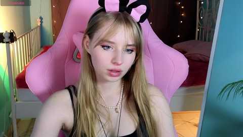 Media: Video of a young Caucasian woman with long blonde hair, wearing a pink bunny headband, black tank top, and necklace, sitting in a pink gaming chair, with a colorful bedroom in the background.