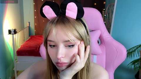 Media: Video of a young, fair-skinned woman with straight blonde hair and large pink mouse ears, resting her head on her hand, in a cozy bedroom with pink furniture and fairy lights.