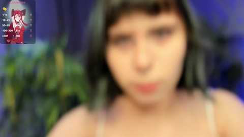 Media: A blurry video of a woman with dark hair and fair skin, wearing a beige top. The background features green foliage and a dark wall.