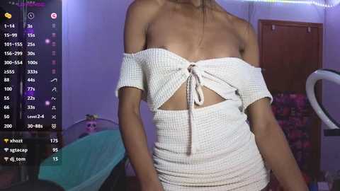 Media: Video of a slender, light-skinned woman in a white, off-the-shoulder, knitted dress with a knot at the waist, standing in a dimly lit room with purple walls, a bed, and a pink chair.