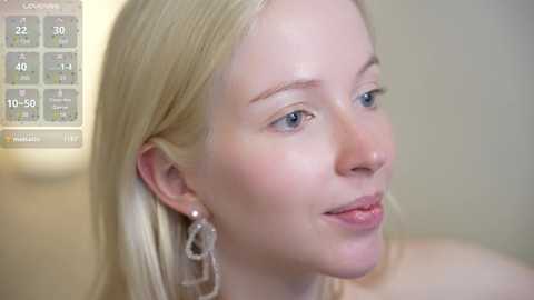 Media: Video of a young, fair-skinned woman with straight blonde hair, wearing silver hoop earrings, looking to the right with a neutral expression. Background includes a blurred thermometer and beige wall.