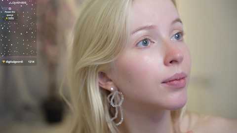 Media: Video of a fair-skinned, blonde-haired woman with blue eyes, wearing large, silver hoop earrings. Background is out of focus, displaying a social media video overlay.