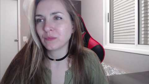 Media: Video of a young, fair-skinned woman with long, straight blonde hair, wearing a green shirt and a black choker, sitting in a modern room with a red and black gaming chair and white blinds.