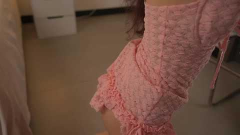 Media: Video of a woman in a pink, ruffled, off-the-shoulder dress standing in a room with beige tiled floors, white filing cabinets, and a white chair.