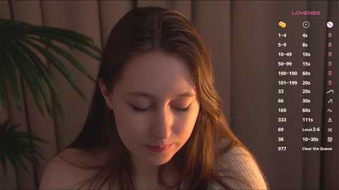 Media: A video of a young woman with long brown hair, fair skin, and closed eyes, wearing a beige sweater, indoors with warm lighting, surrounded by a green plant and wooden background.