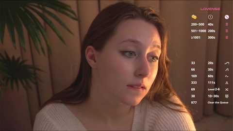 Media: Video of a young woman with fair skin, light brown hair, and green eyes, wearing a beige sweater, looking introspective. Background features a wooden fence and a potted plant.