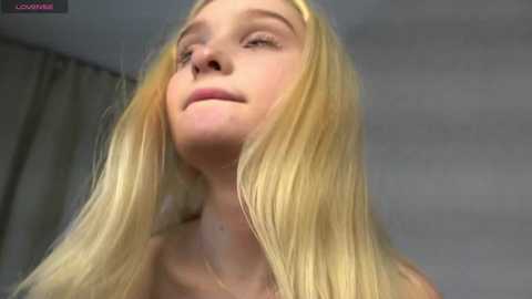 Media: Video of a young Caucasian woman with long, straight blonde hair, fair skin, and a neutral expression. She appears topless, with the background featuring light-colored curtains and a soft-focus, gray wall.