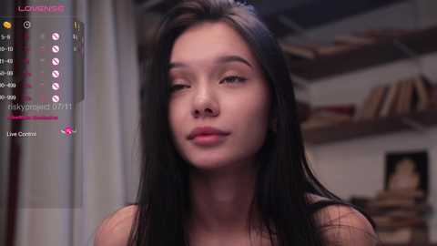 Media: Video of an Asian woman with long black hair, wearing a tank top, in a dimly lit room with shelves in the background.