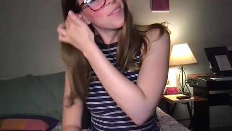 Media: Video of a young woman with fair skin and long brown hair, wearing glasses, adjusting her hair in a living room with a lamp, gray sofa, and books.